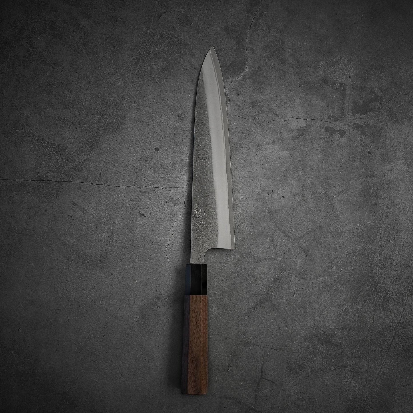 A Yoshikane nashiji shirogami#2 gyuto 240mm chef’s knife by Yoshikane, featuring a wooden handle, rests vertically on a textured gray concrete surface with its stainless steel blade pointing upward.