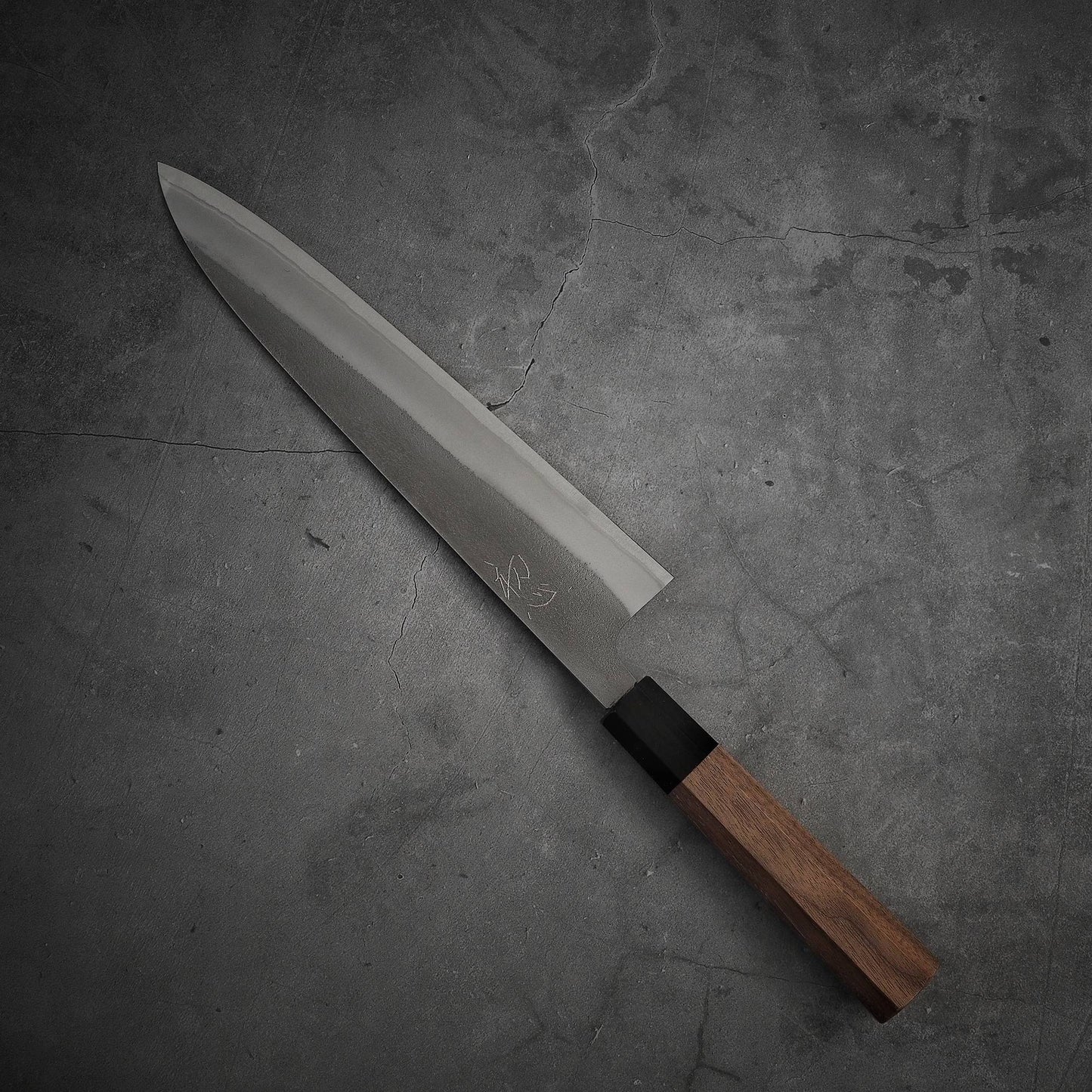 The Yoshikane nashiji shirogami#2 gyuto 240mm, featuring a long, pointed stainless steel blade and a wooden handle, rests on a textured gray concrete surface.