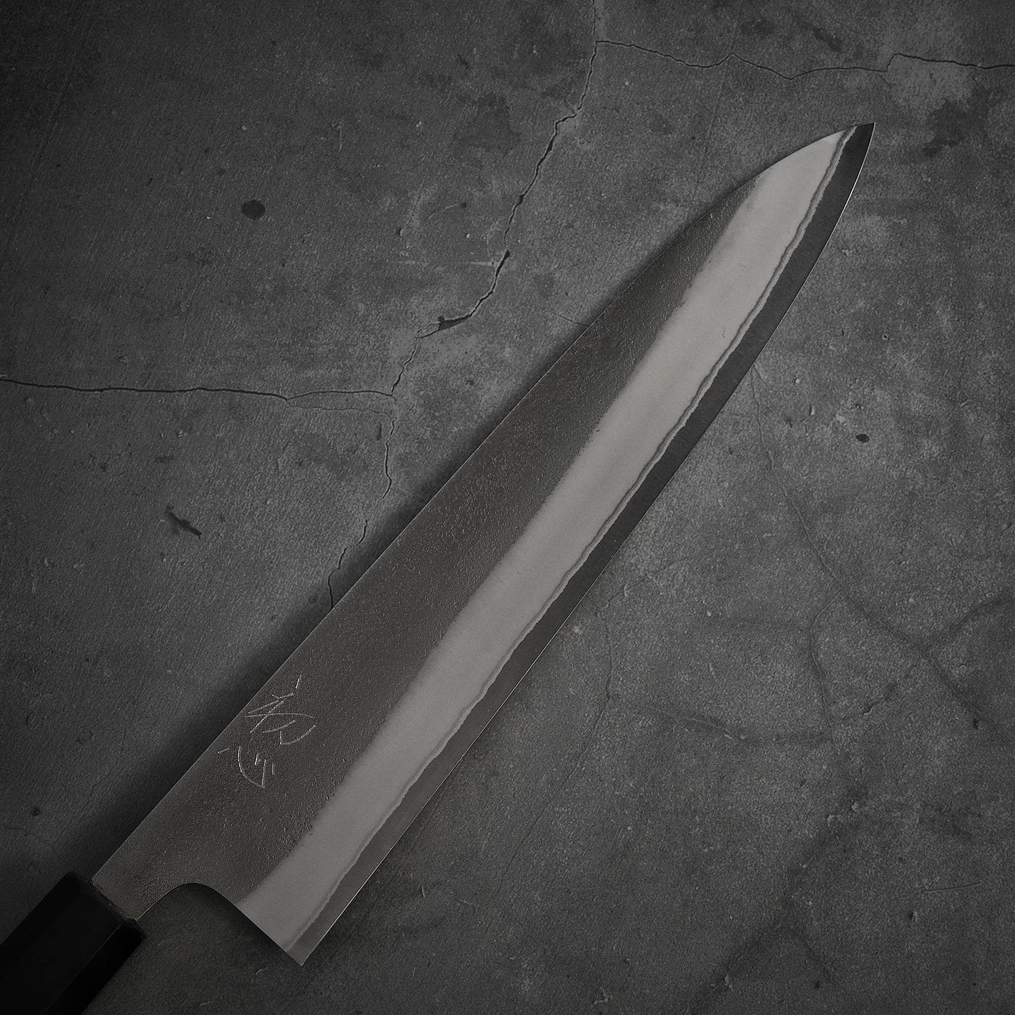 The Yoshikane nashiji shirogami#2 gyuto 240mm features engraved symbols and rests on a cracked, textured gray surface.