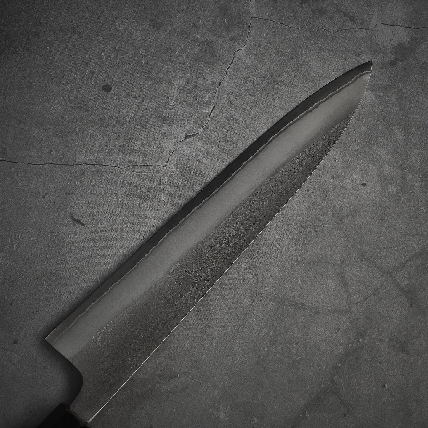 A Yoshikane nashiji shirogami#2 gyuto 240mm knife with a sharp, patterned blade rests on cracked gray concrete, showcasing the Yoshikane brands expert steel craftsmanship.