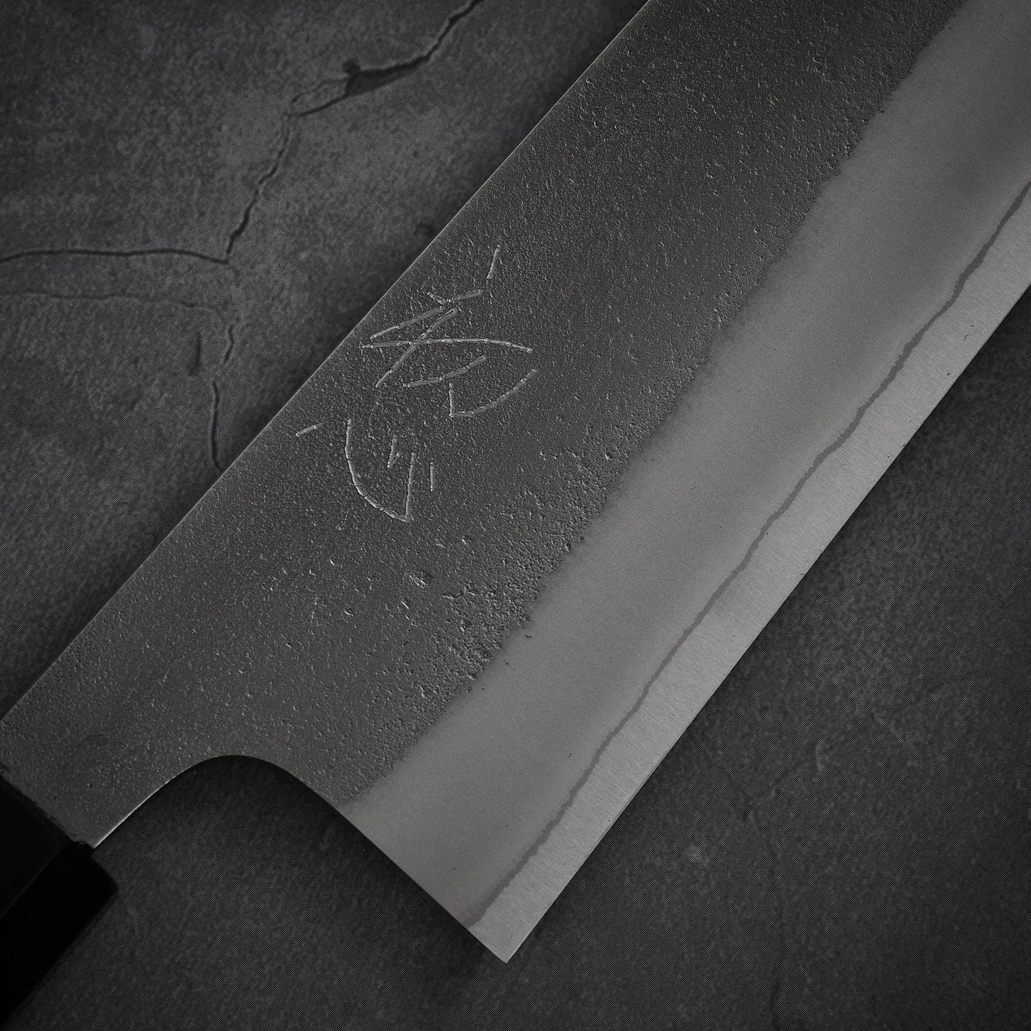 Close-up view of the Yoshikane nashiji shirogami#2 gyuto 240mm by Yoshikane, featuring kanji on its shirogami#2 steel blade with a textured, polished finish against a dark cracked background.