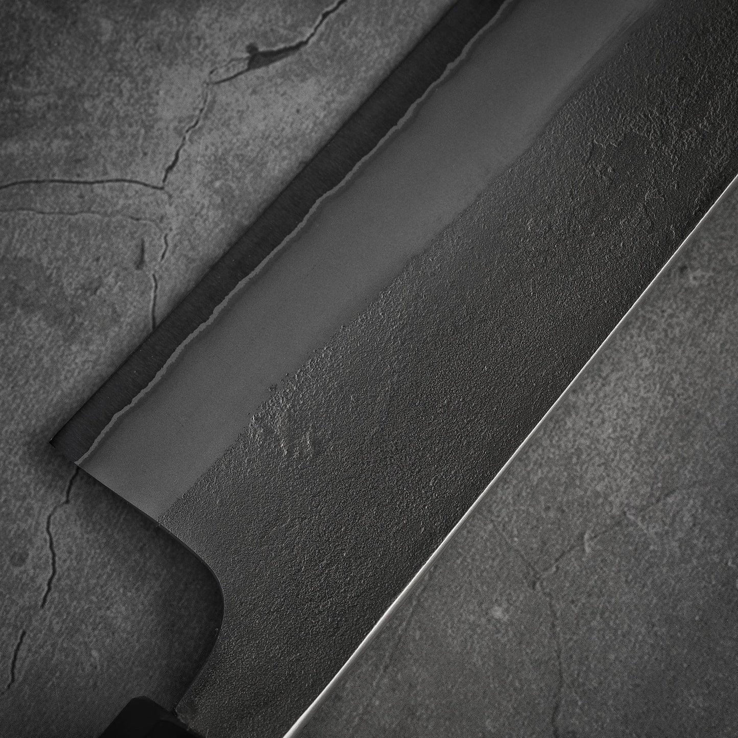Close-up of the Yoshikane nashiji shirogami#2 gyuto 240mm blade by Yoshikane on a cracked gray surface, highlighting the matte shirogami#2 steel grain and edge, with part of the handle visible in the lower left corner.