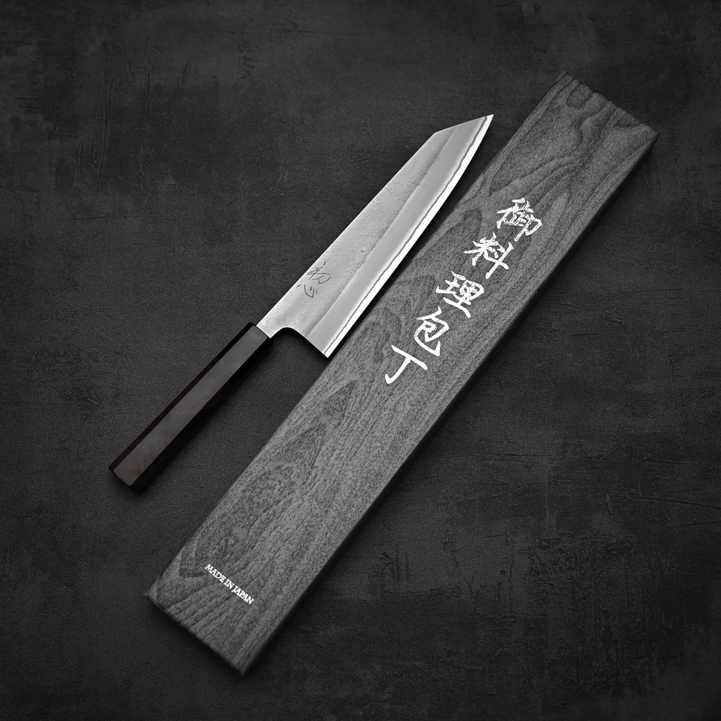 The Yoshikane Hatsukokoro x Yoshikane nashiji shirogami#2 kiritsuke gyuto 210mm, featuring a dark handle, rests by a gray wooden case with white Japanese characters on a textured dark background. This elegant blade is crafted from shirogami#2 steel.