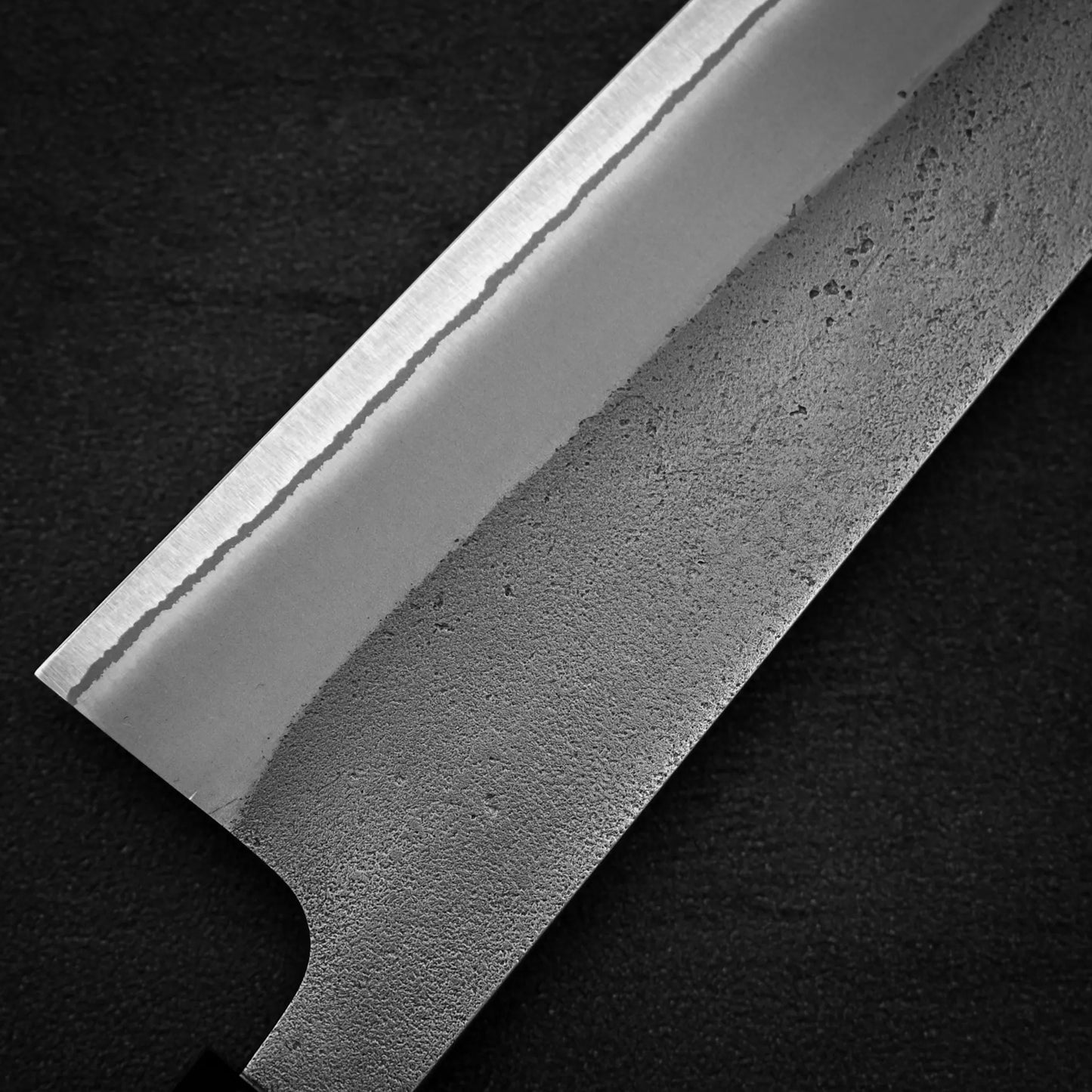 Close-up of the Yoshikane Hatsukokoro x Yoshikane nashiji shirogami#2 kiritsuke gyuto 210mm blade, showcasing its nashiji textured finish, distinct hamon line, and razor-sharp edge against a dark background.