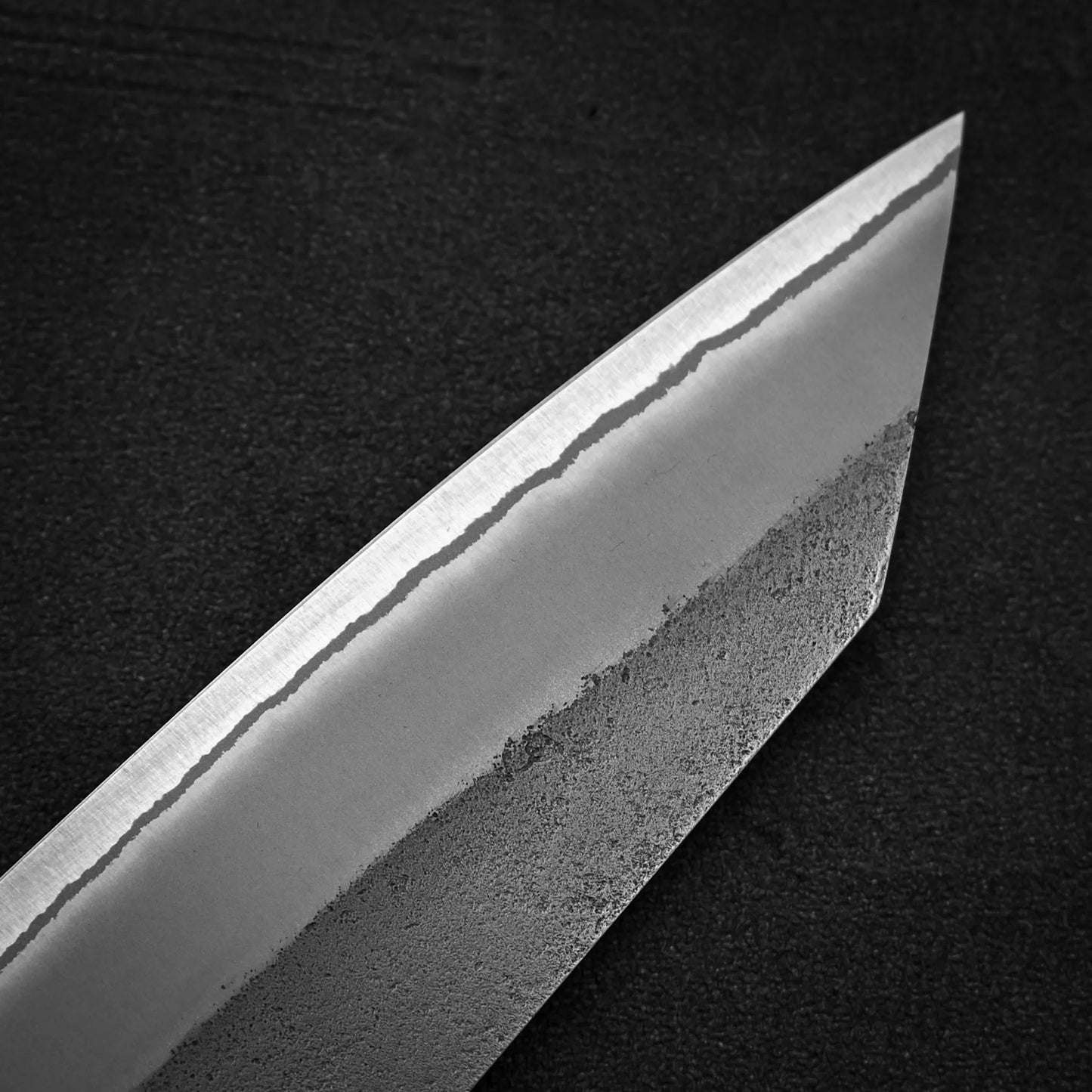 Close-up of the pointed tip of the Yoshikane Hatsukokoro x Yoshikane nashiji shirogami#2 kiritsuke gyuto 210mm blade, showing its wavy nashiji pattern, polished upper part, and textured lower edge against a dark background.