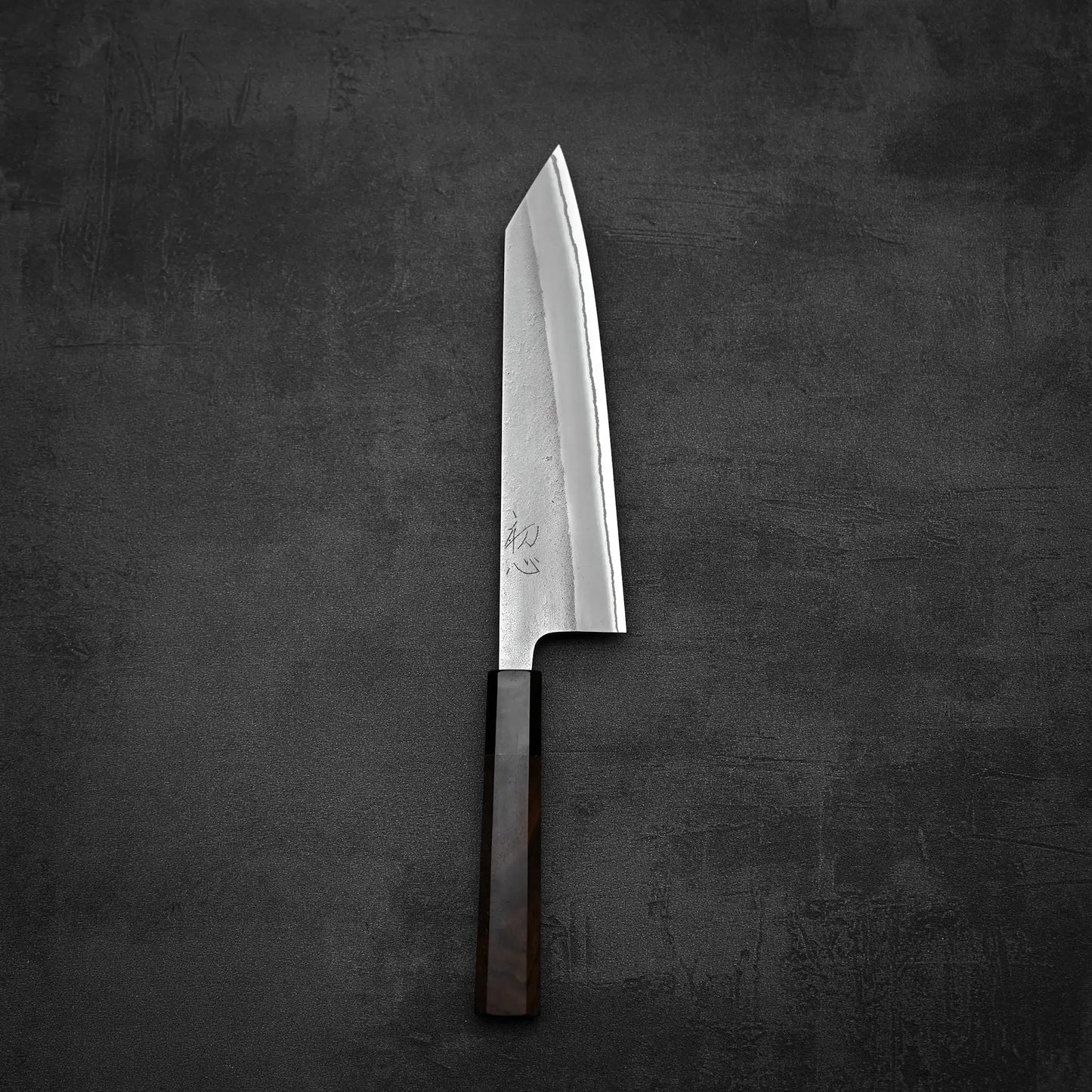 The Yoshikane Hatsukokoro x Yoshikane nashiji shirogami#2 kiritsuke gyuto 210mm chefs knife rests on a dark, textured surface, showcasing its rectangular wooden handle and angled, sharp blade.