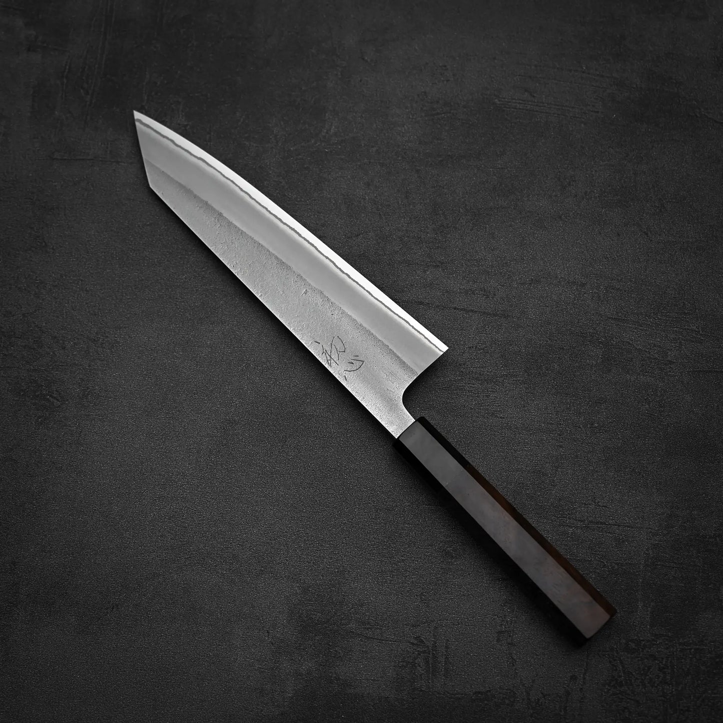 The Yoshikane Hatsukokoro x Yoshikane nashiji shirogami#2 kiritsuke gyuto 210mm, featuring a sharp pointed blade and dark wooden handle, rests on a black textured surface.