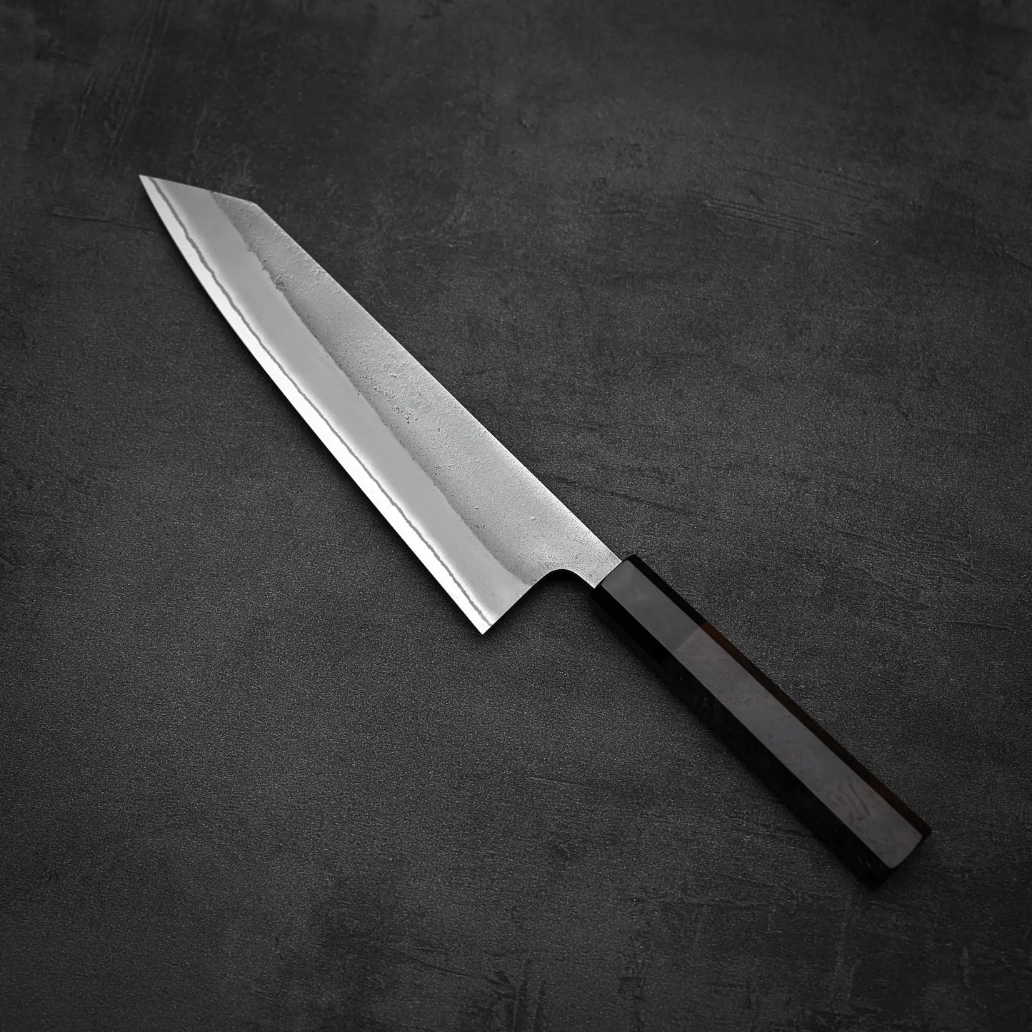 The Yoshikane Hatsukokoro x Yoshikane nashiji shirogami#2 kiritsuke gyuto 210mm, featuring a pointed blade and dark handle, rests on a dark, textured surface.