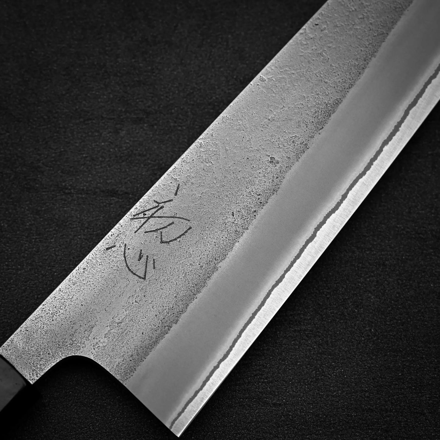 Close-up of a Yoshikane Hatsukokoro x Yoshikane nashiji shirogami#2 kiritsuke gyuto 210mm, featuring a textured blade with kanji engraving. The polished edge contrasts with the matte upper section against a dark background.