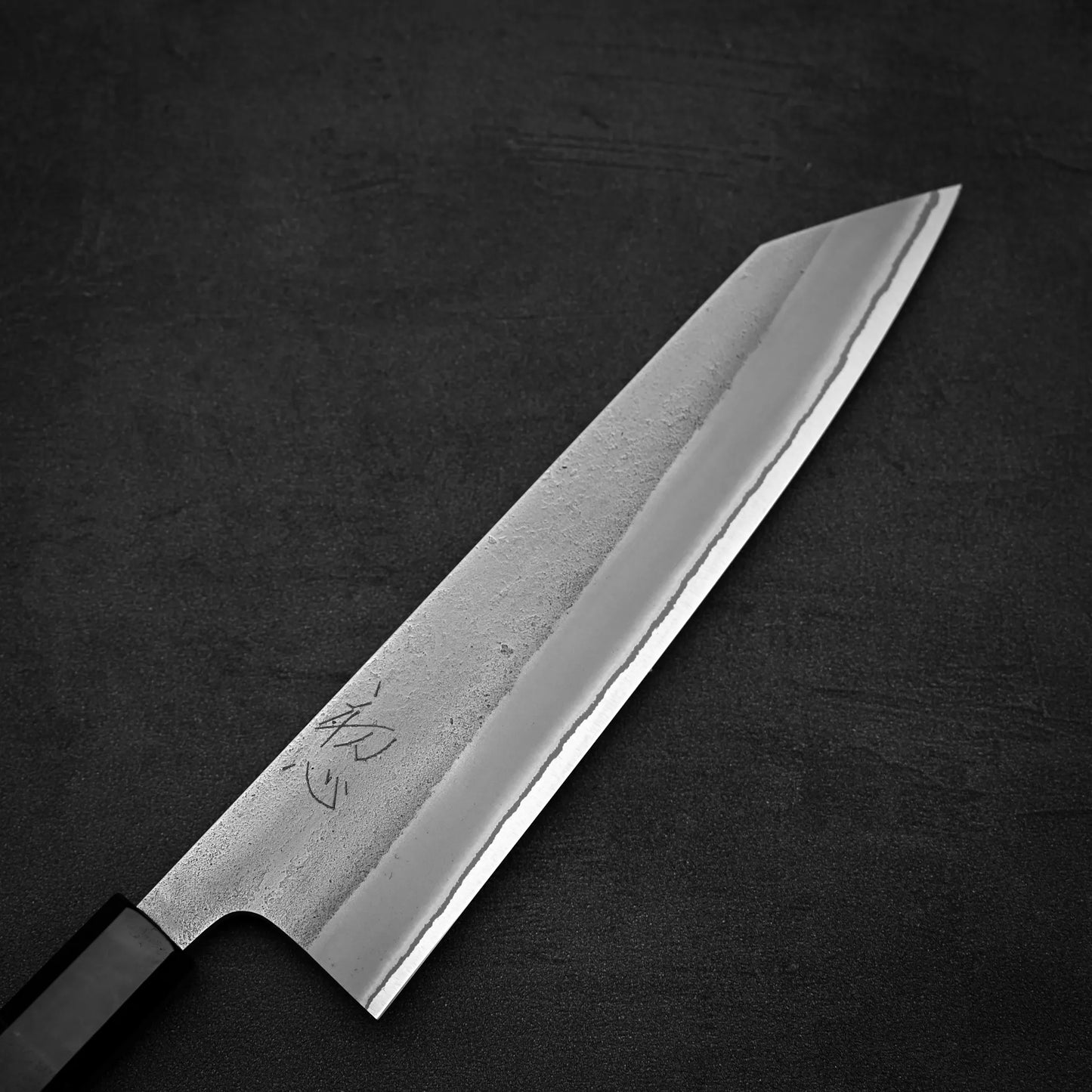 The Yoshikane Hatsukokoro x Yoshikane nashiji shirogami#2 kiritsuke gyuto 210mm chef’s knife features a sharp, angled blade with distinctive engravings and a black handle, displayed on a dark textured surface.