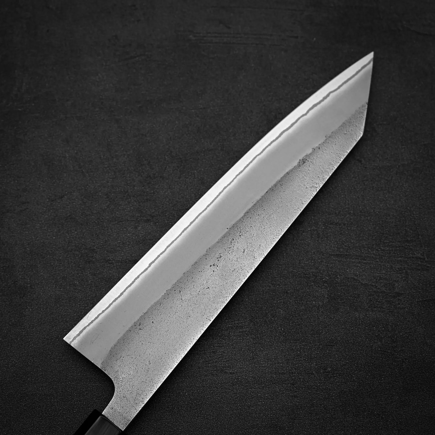A close-up of the Hatsukokoro x Yoshikane nashiji shirogami#2 kiritsuke gyuto 210mm by Yoshikane, featuring a sharp, angled blade made from shirogami#2 steel, set against a dark, textured background.