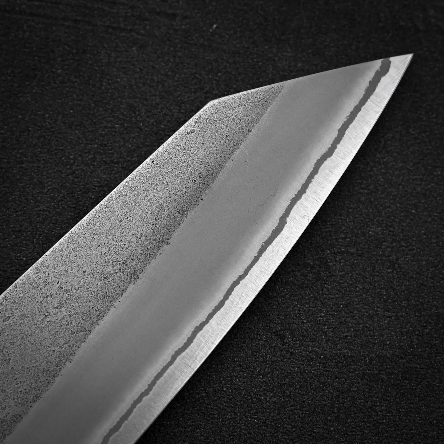 Close-up of the Yoshikane Hatsukokoro x Yoshikane nashiji shirogami#2 kiritsuke gyuto 210mm blade, featuring a visible hamon that highlights the contrast between its polished edge and textured upper section against a dark background.