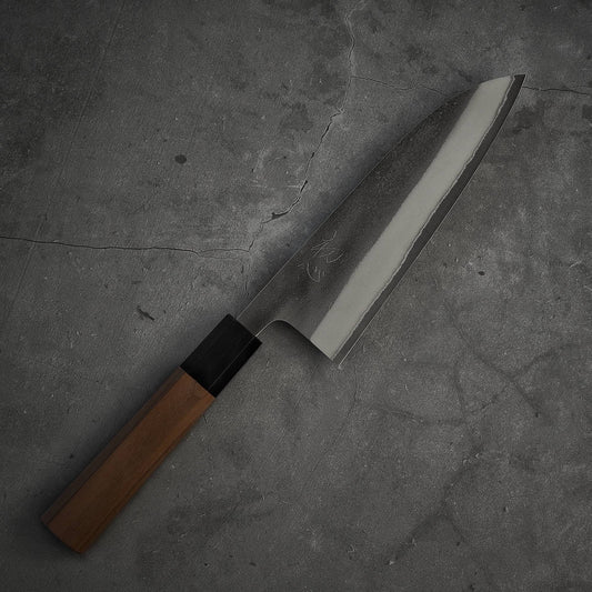 The Yoshikane nashiji shirogami#2 santoku 165mm knife by Yoshikane, with a wooden handle and sharp blade, rests on a gray textured stone surface.