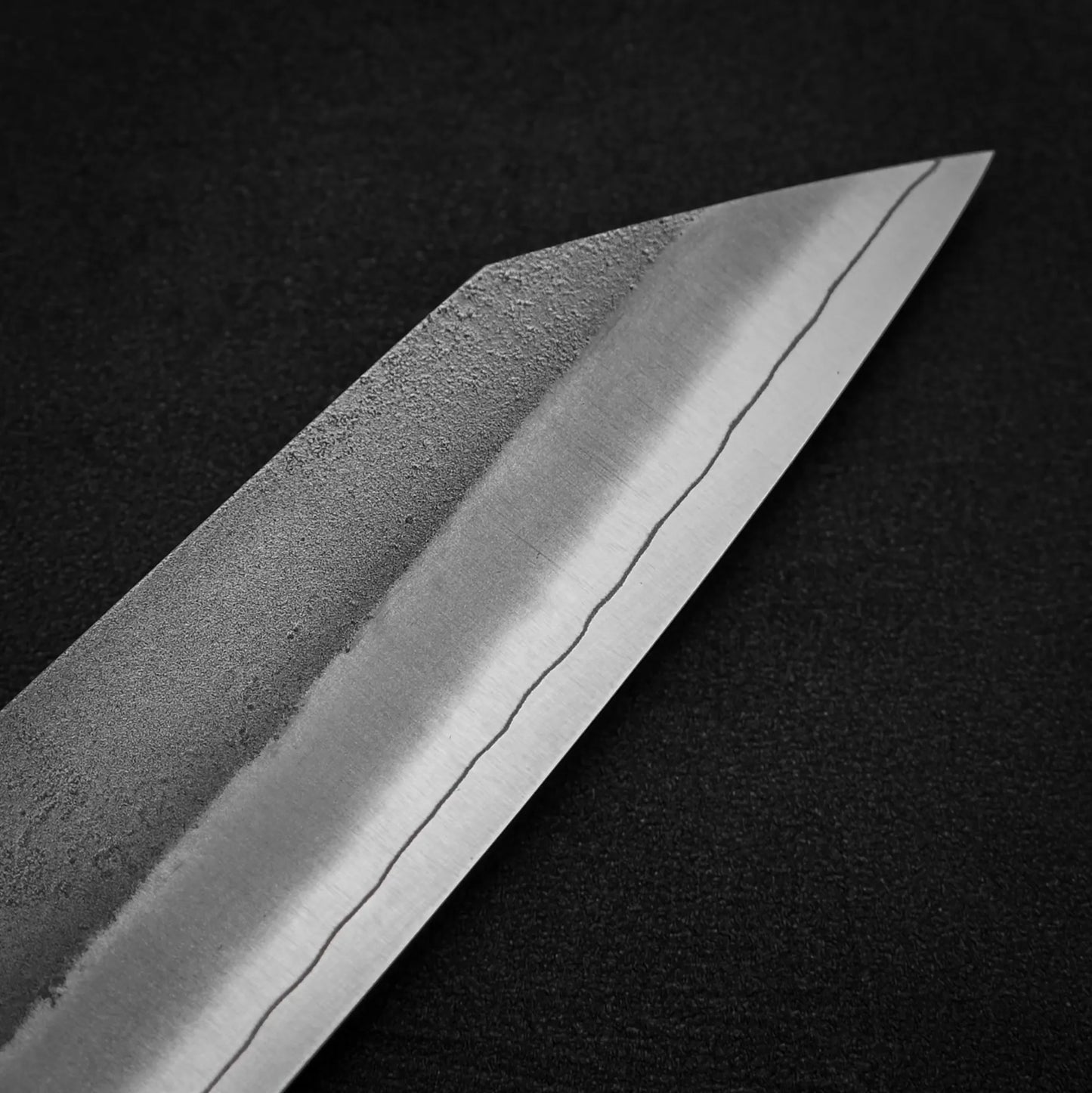 Close-up of the Hatsukokoro x Yoshikane nashiji SKD kiritsuke gyuto 210mm blade by Yoshikane, featuring polished SKD steel and a visible hamon, set against a dark textured background.
