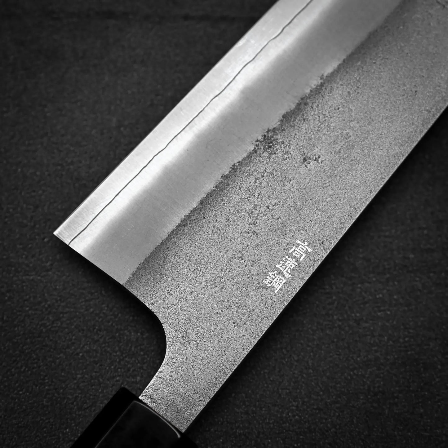 Close-up of the Yoshikane Hatsukokoro x Yoshikane nashiji SKD kiritsuke gyuto 210mm blade on a dark background, highlighting its wavy SKD steel pattern and engraved Japanese characters near the edge.
