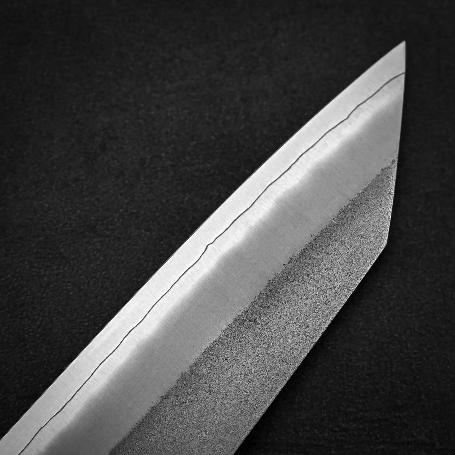 Close-up of the Hatsukokoro x Yoshikane nashiji SKD kiritsuke gyuto 210mm blade tip, showing its wavy pattern and SKD steel, by Yoshikane, photographed against a dark background.