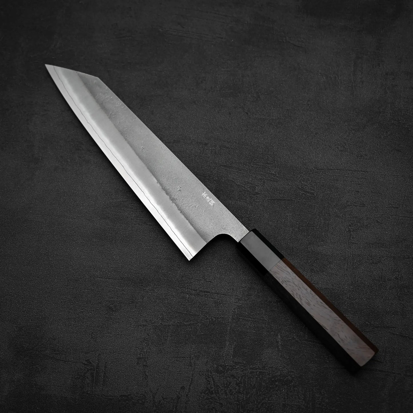 The Yoshikane Hatsukokoro x Yoshikane nashiji SKD kiritsuke gyuto 210mm, with a sharp, angled blade and dark wooden handle, rests on a textured black surface.