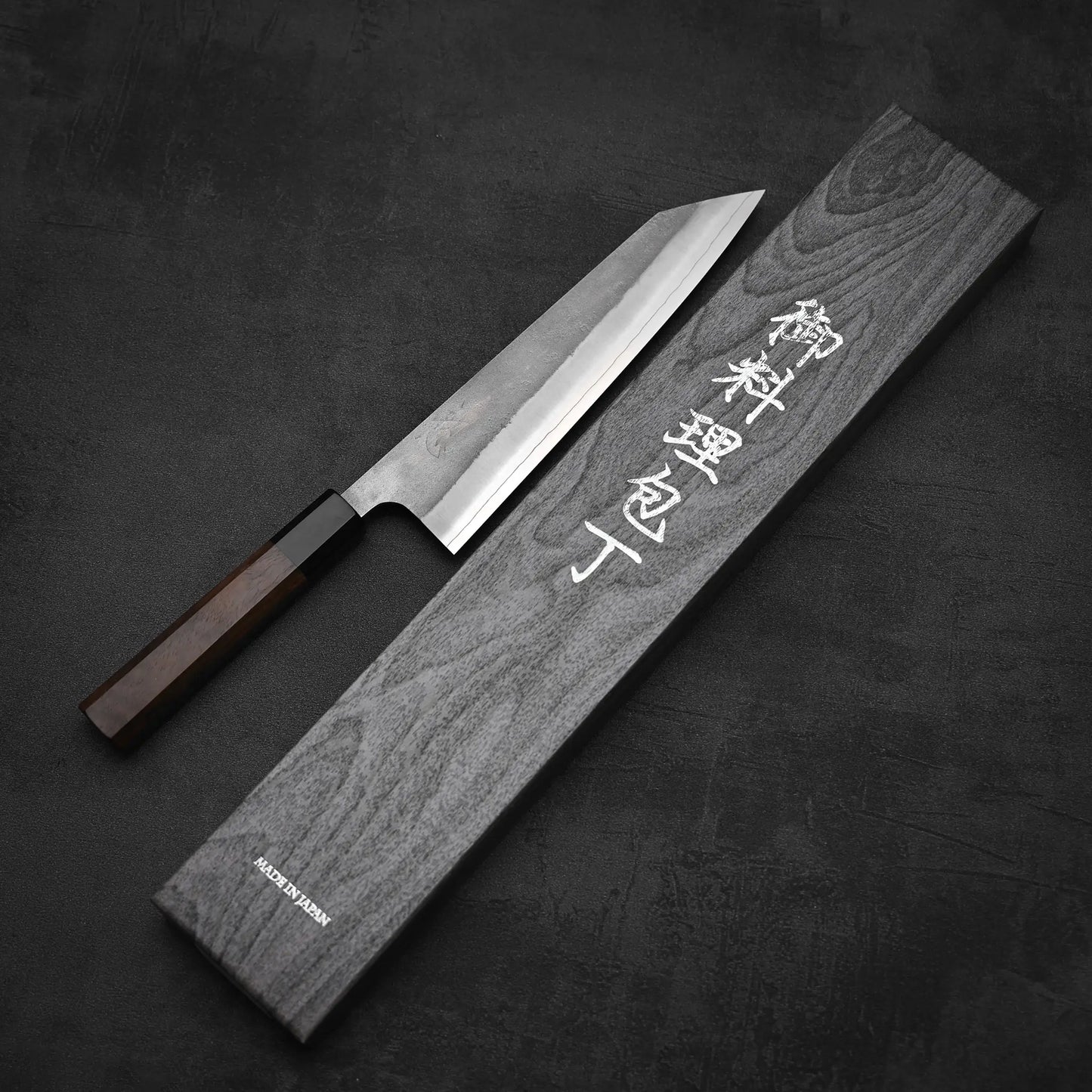 A Yoshikane Hatsukokoro x Yoshikane nashiji SKD kiritsuke gyuto 210mm with a dark wooden handle rests beside a rectangular box with Japanese characters on the lid, all placed on a dark textured surface.