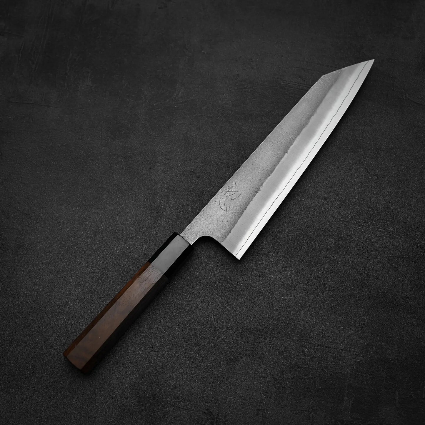 The Yoshikane Hatsukokoro x Yoshikane nashiji SKD kiritsuke gyuto 210mm, featuring a sharp angular blade and dark wooden handle, rests on a textured black surface.