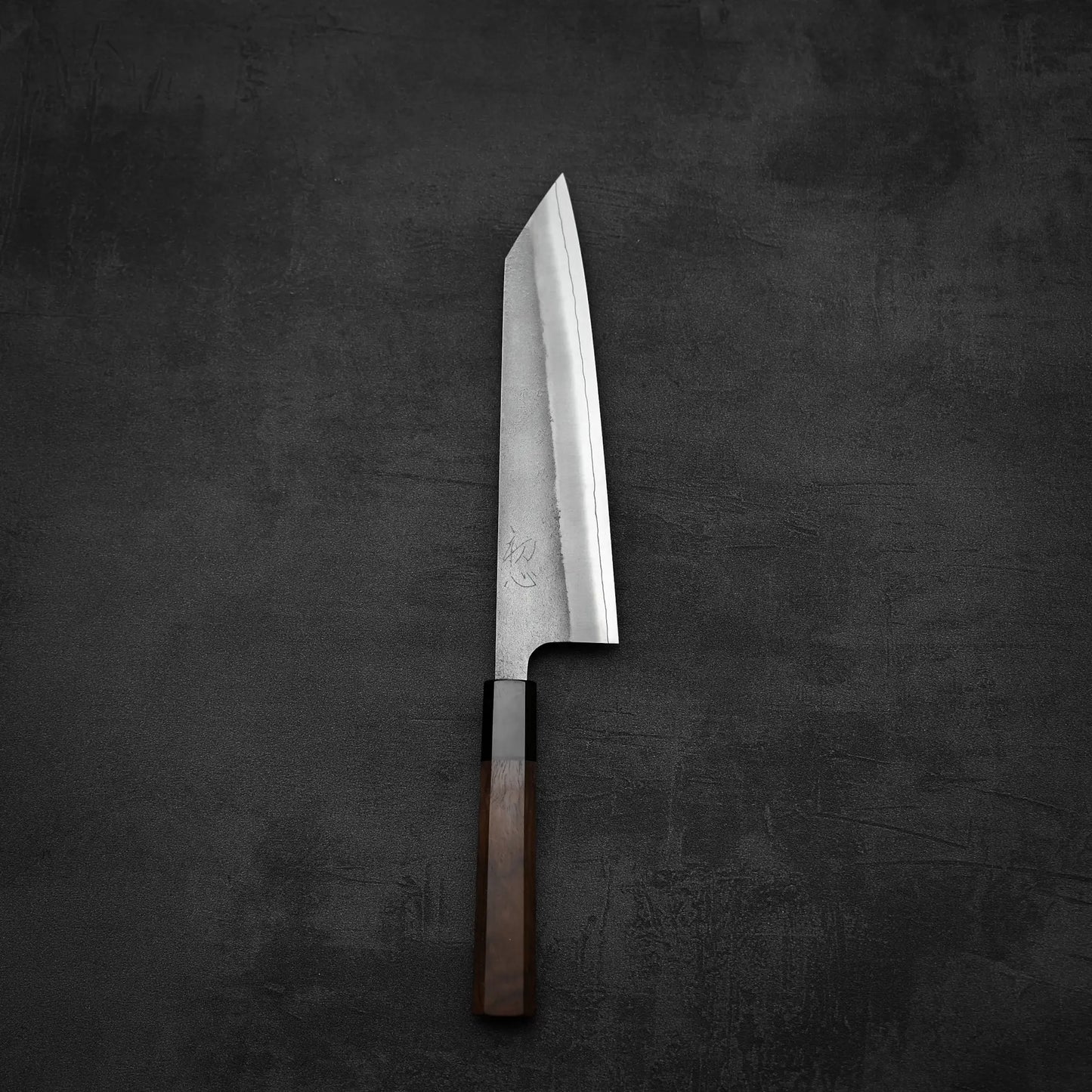 A Yoshikane Hatsukokoro x Yoshikane nashiji SKD kiritsuke gyuto 210mm with a wooden handle and pointed tip is placed on a dark textured surface.