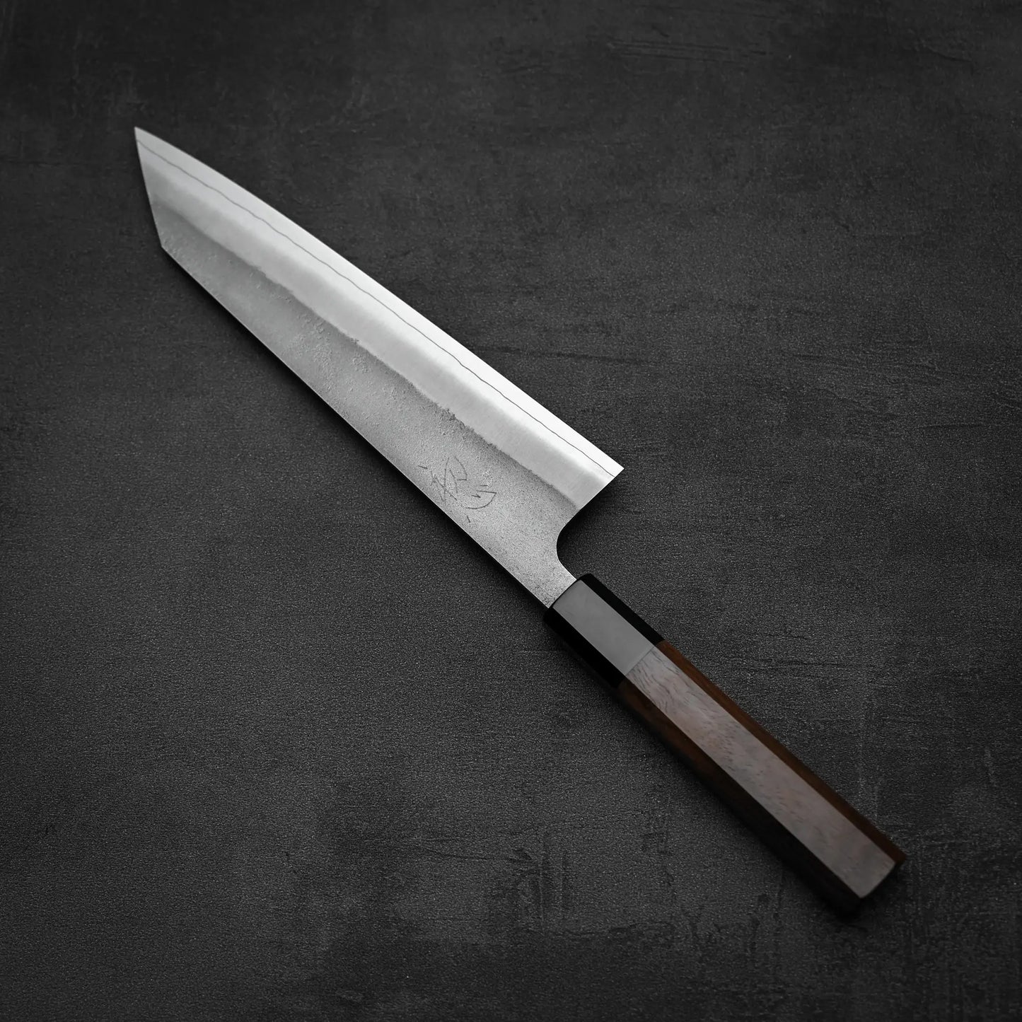 A Yoshikane Hatsukokoro x Yoshikane nashiji SKD kiritsuke gyuto 210mm knife with a sharp blade and dark wooden handle rests on a dark, textured surface.