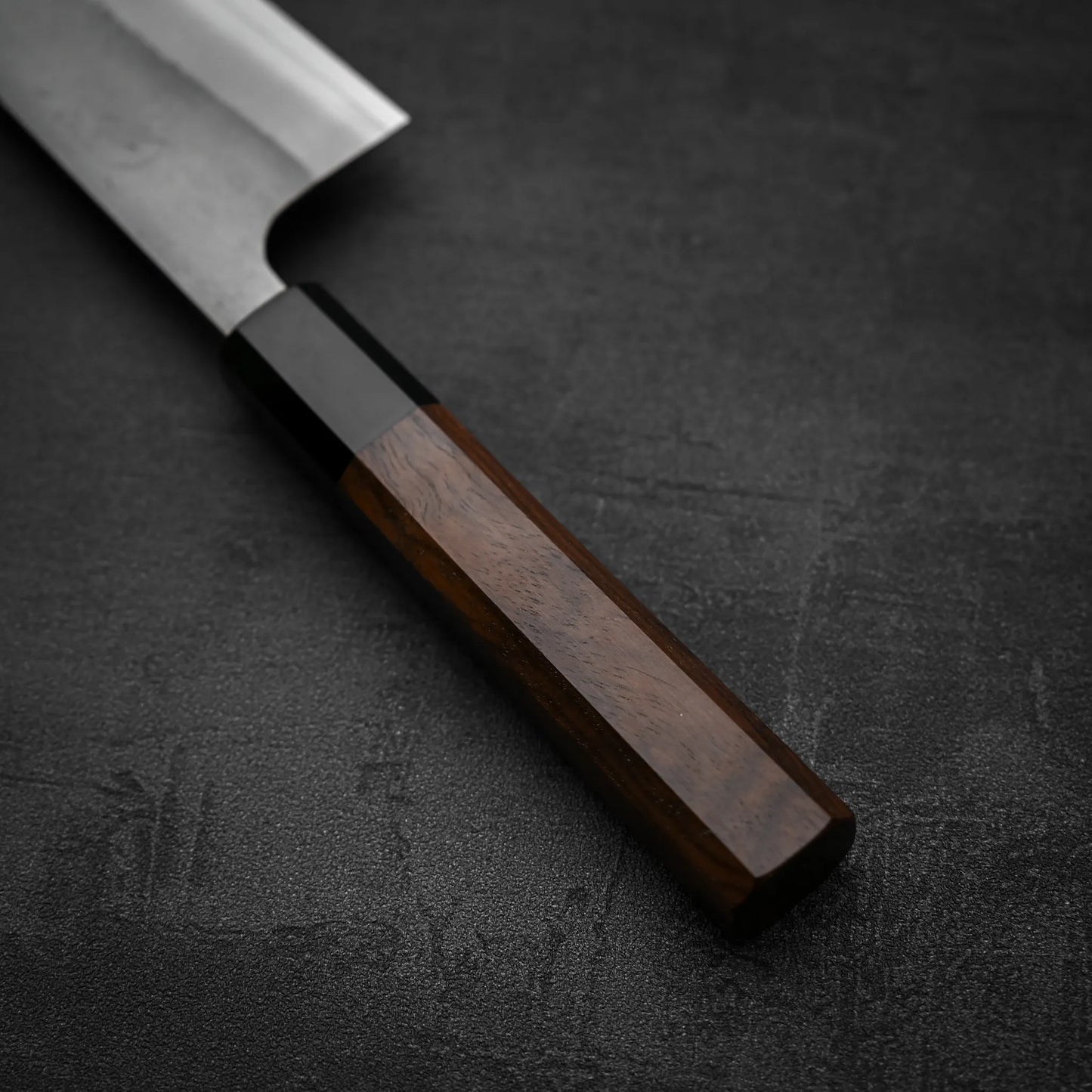 A close-up of the Yoshikane Hatsukokoro x Yoshikane nashiji SKD kiritsuke gyuto 210mm knife with a wooden handle, resting on a dark textured surface.