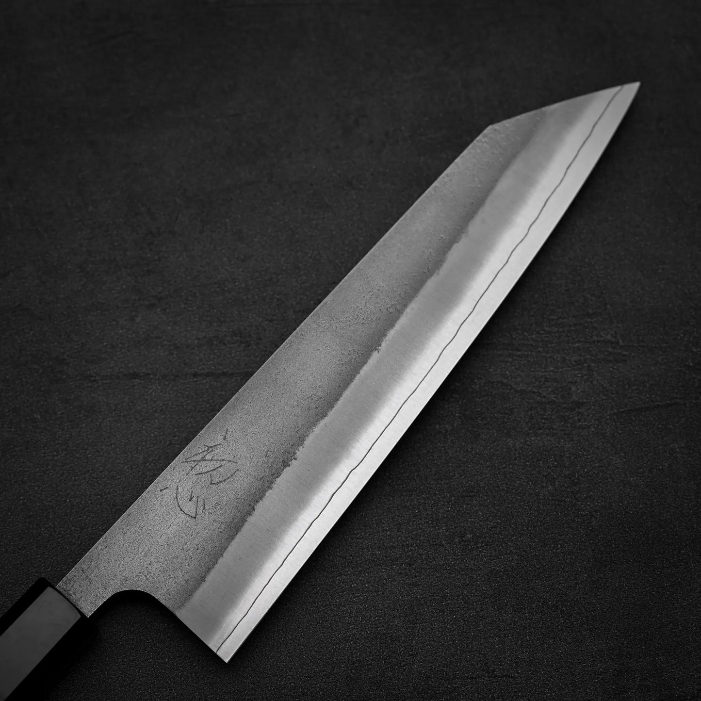 A close-up of the Yoshikane Hatsukokoro x Yoshikane nashiji SKD kiritsuke gyuto 210mm knife, showcasing its sharp, pointed SKD steel blade with a visible hamon pattern against a dark textured background.