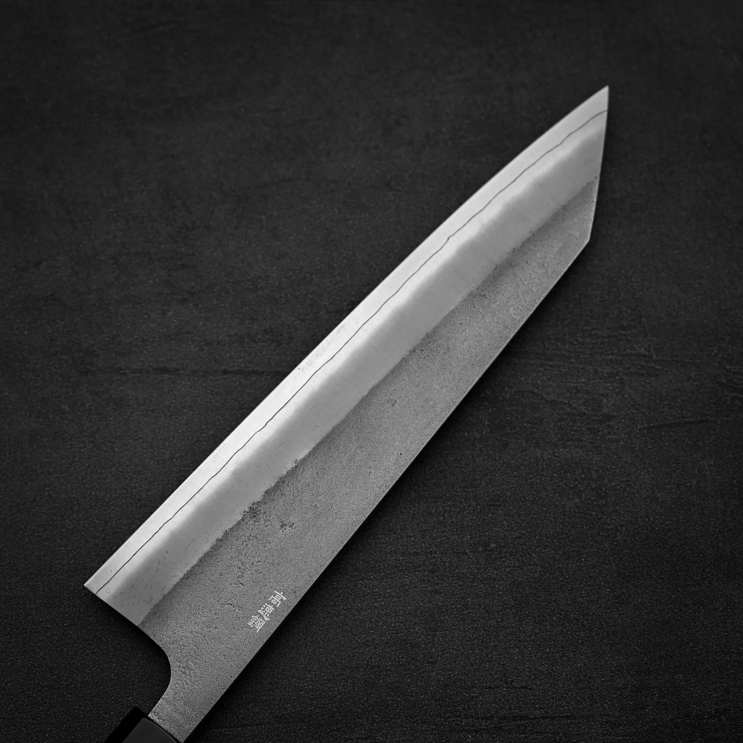 A close-up of the Yoshikane Hatsukokoro x Yoshikane nashiji SKD kiritsuke gyuto 210mm knife, showing its sharp, angled blade with a wavy pattern, crafted in Japan from SKD steel, set against a dark textured background.