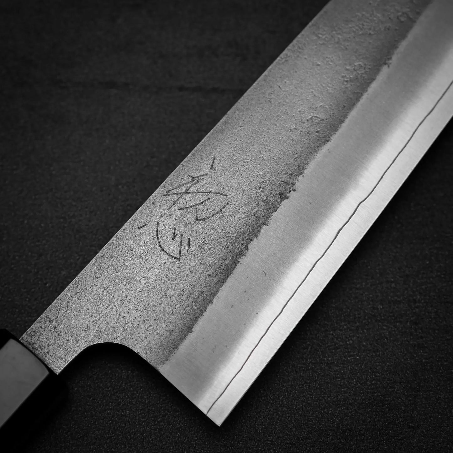 Close-up of a Yoshikane Hatsukokoro x Yoshikane nashiji SKD kiritsuke gyuto 210mm blade, featuring visible kanji engraving and a distinct wavy SKD steel edge, set against a dark background.