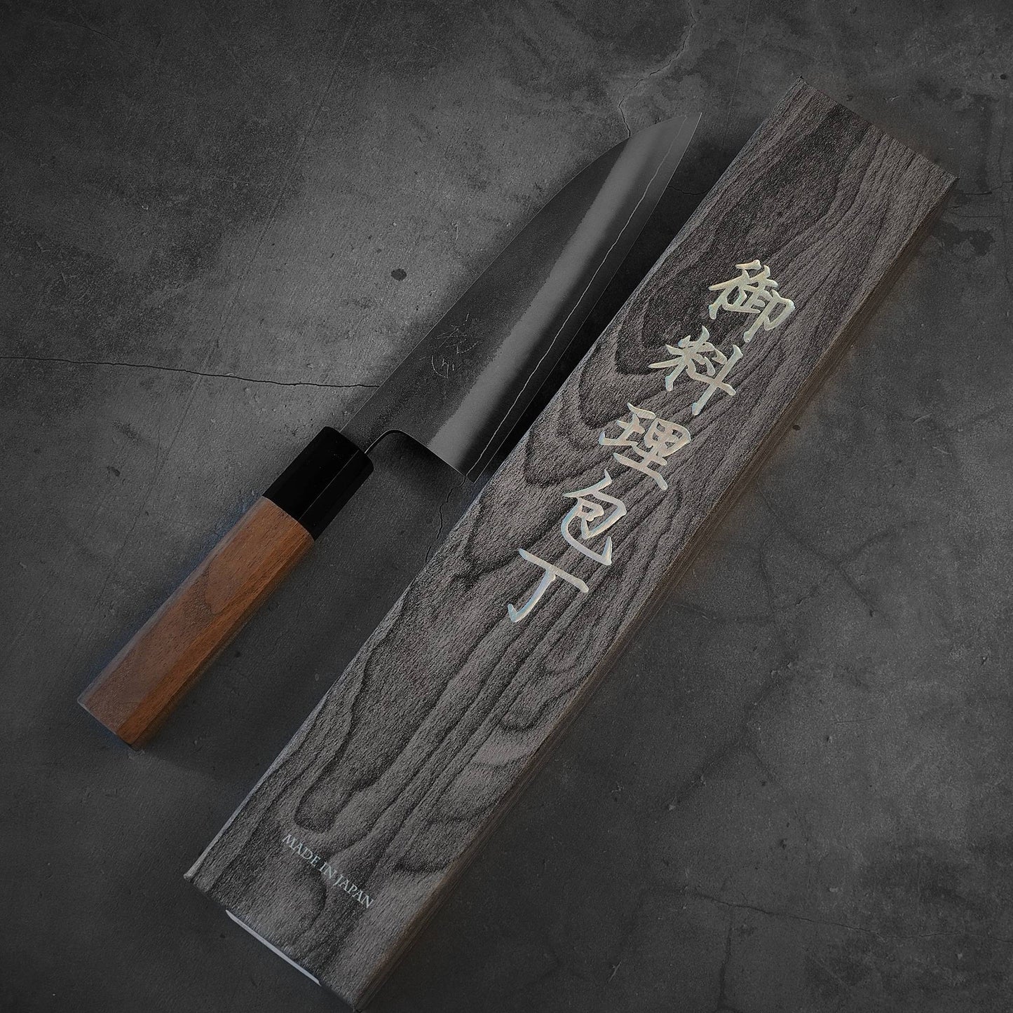 A Yoshikane nashiji SKD santoku 165mm kitchen knife by Yoshikane, featuring a wooden handle, rests beside its engraved wooden box on a dark, textured surface.