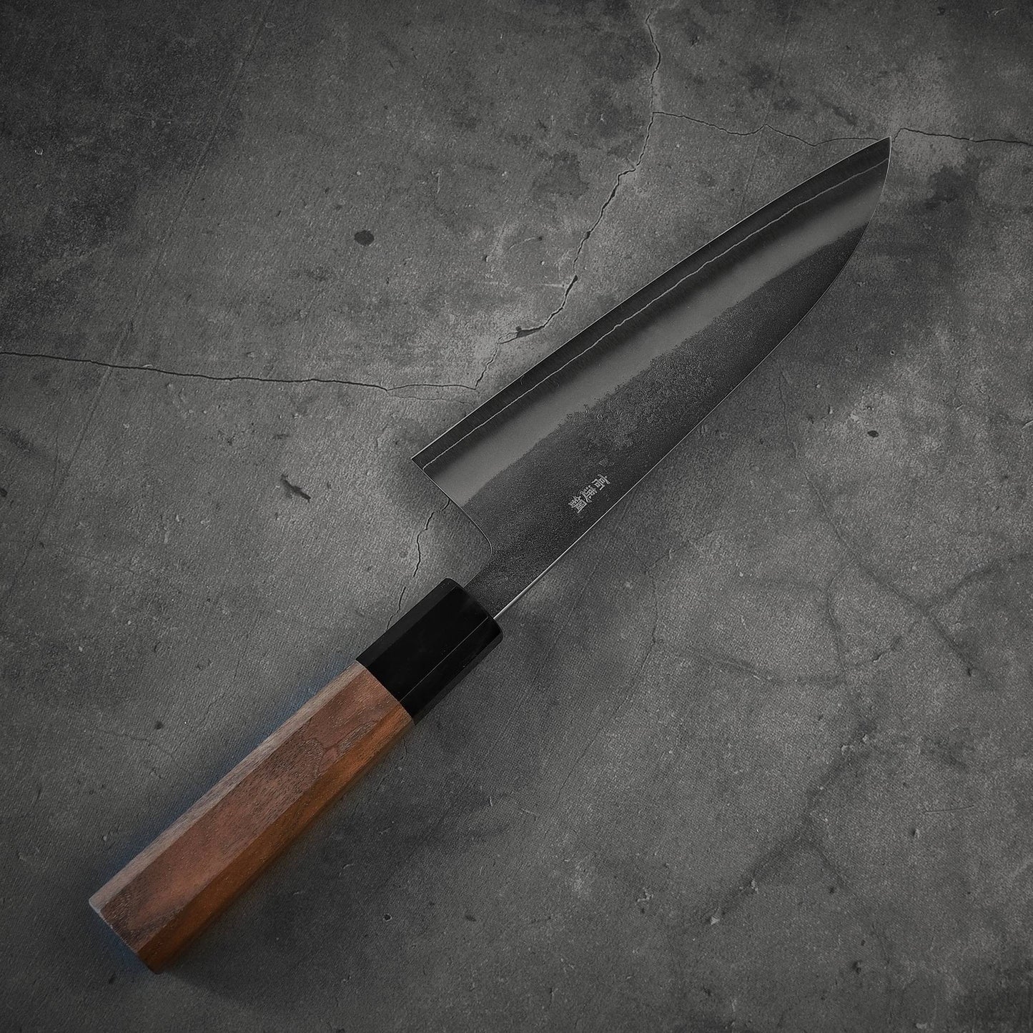 The Yoshikane nashiji SKD santoku 165mm, a Japanese-style kitchen knife with a dark SKD steel blade and wooden handle, rests on a cracked, gray stone surface.