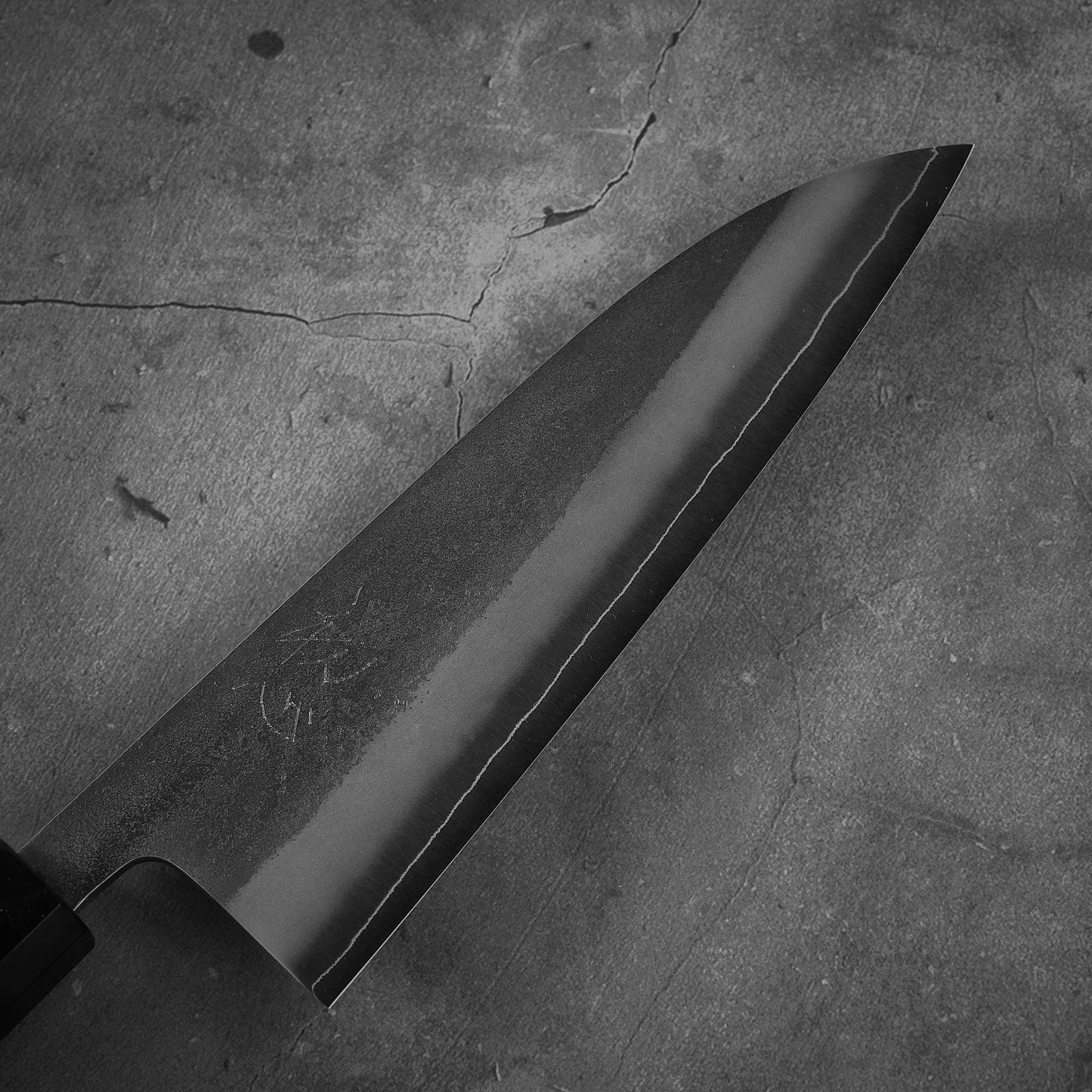 A close-up of a Yoshikane nashiji SKD santoku 165mm chef’s knife with a dark SKD steel blade, resting on rough, cracked concrete.