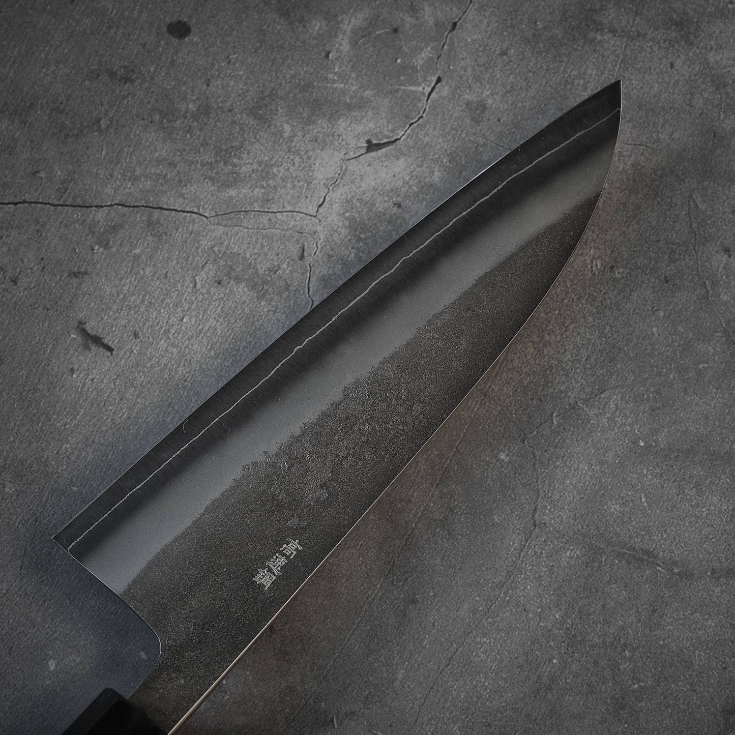 A close-up of the Yoshikane nashiji SKD santoku 165mm chef’s knife by Yoshikane, featuring Japanese characters on its dark SKD steel blade, displayed on a gray cracked stone surface.