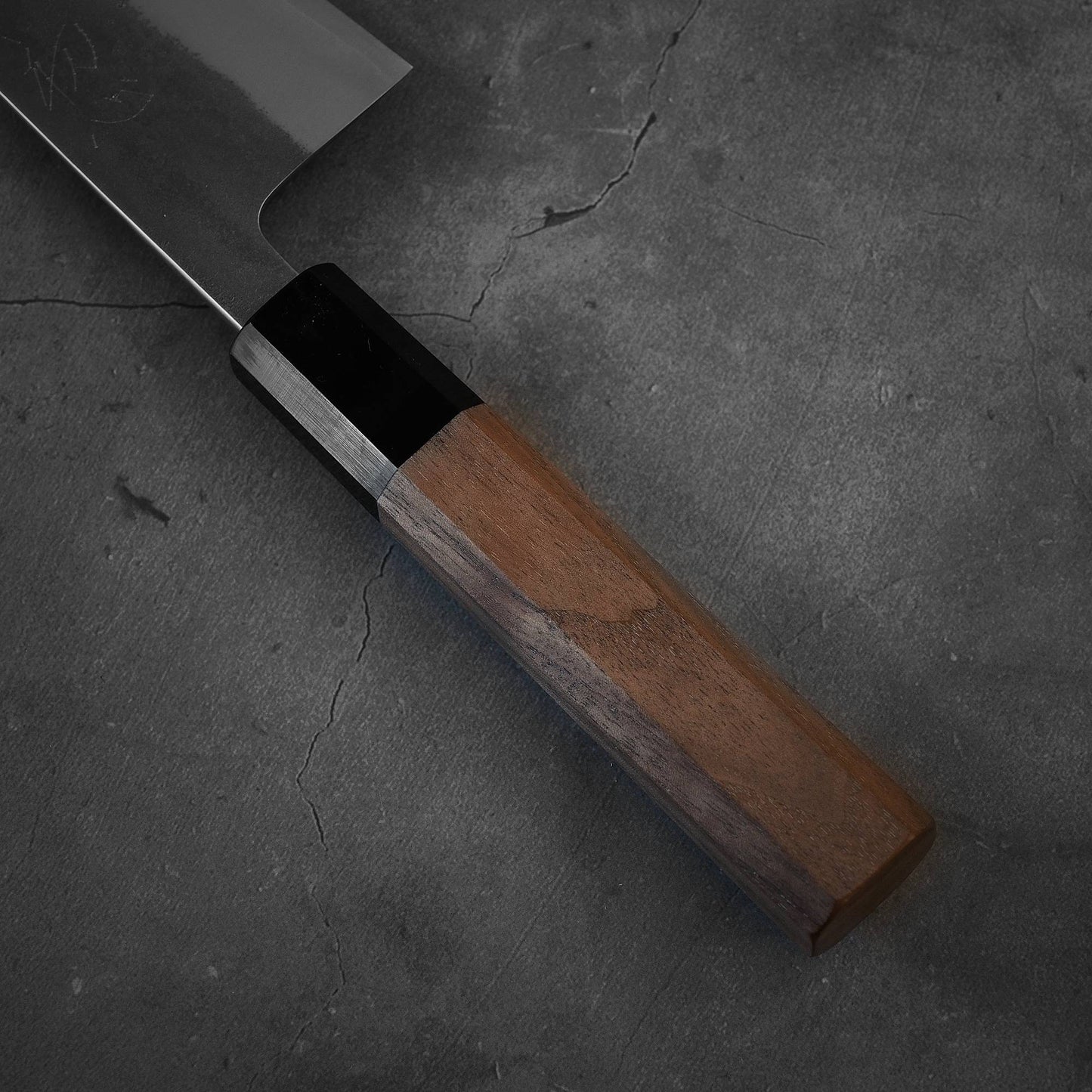 A close-up of a Yoshikane nashiji SKD santoku 165mm kitchen knife by Yoshikane, featuring a wooden handle and SKD steel blade, resting on a dark, cracked concrete surface.