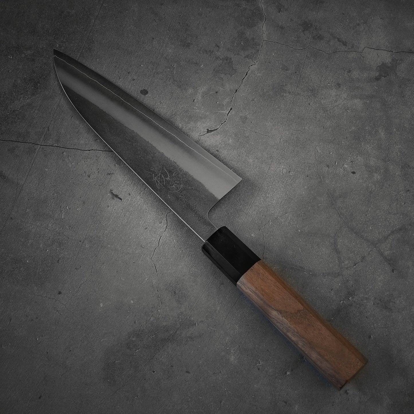 A Yoshikane nashiji SKD santoku 165mm chefs knife by Yoshikane with a dark SKD steel blade and wooden handle rests on a textured gray concrete surface.