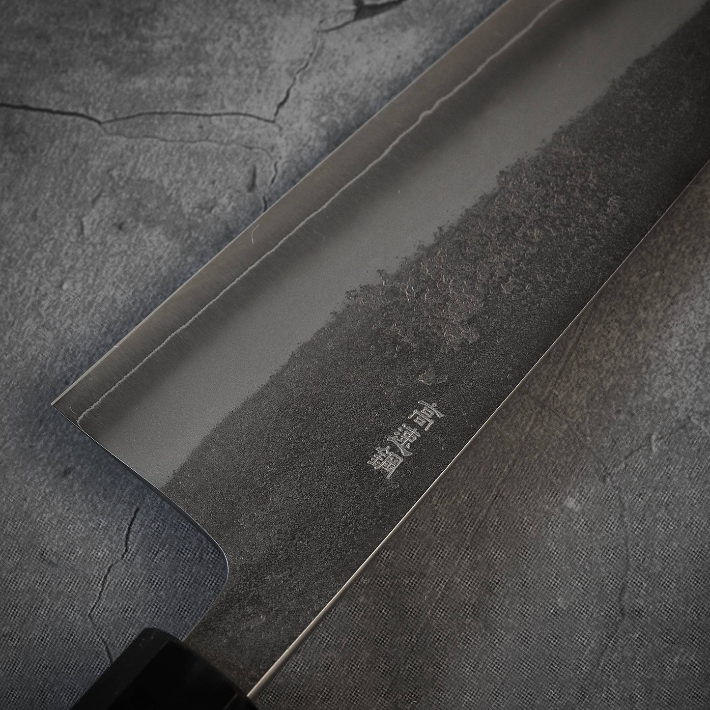 A close-up of a Yoshikane nashiji SKD santoku 165mm Japanese kitchen knife, featuring kanji on the SKD steel blade, resting on a textured cracked gray surface.