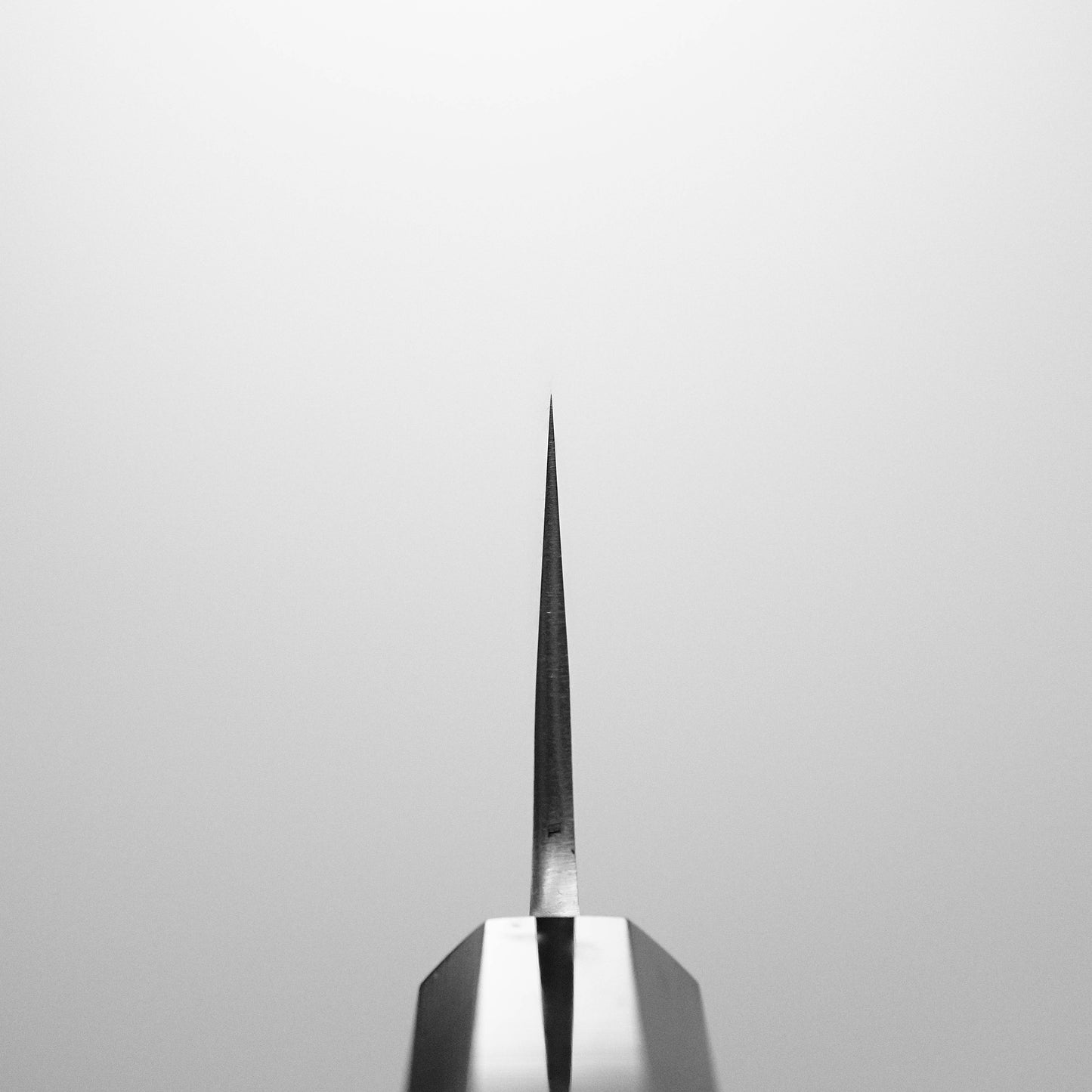 Close-up of the pointed tip of a Yoshikane nashiji SKD santoku 165mm blade, positioned vertically against a plain, light background. The sharp edge is in focus at the center. Brand: Yoshikane.