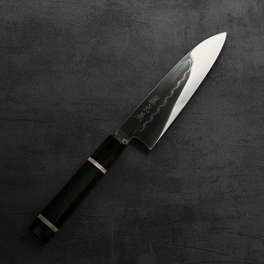 The Yoshikazu Ikeda shirogami#2 mizuhonyaki santoku 165mm, with a dark handle and shiny, engraved blade, rests on a dark textured surface.