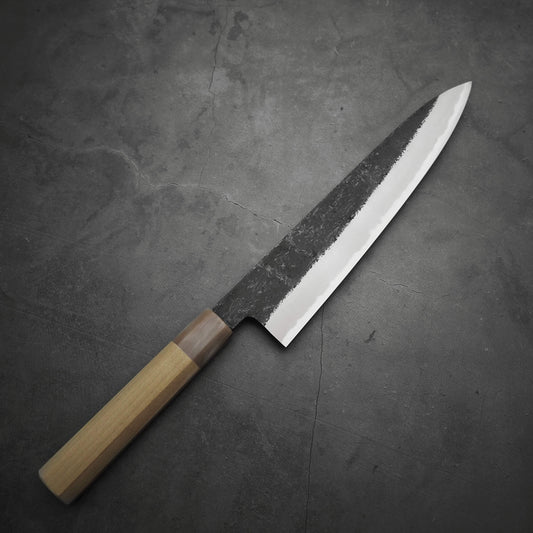 A Yoshikazu Tanaka kurouchi AS gyuto 240mm with a wooden handle and an Aogami Super blade—partially polished, partially textured—rests on a dark, stone-like surface.