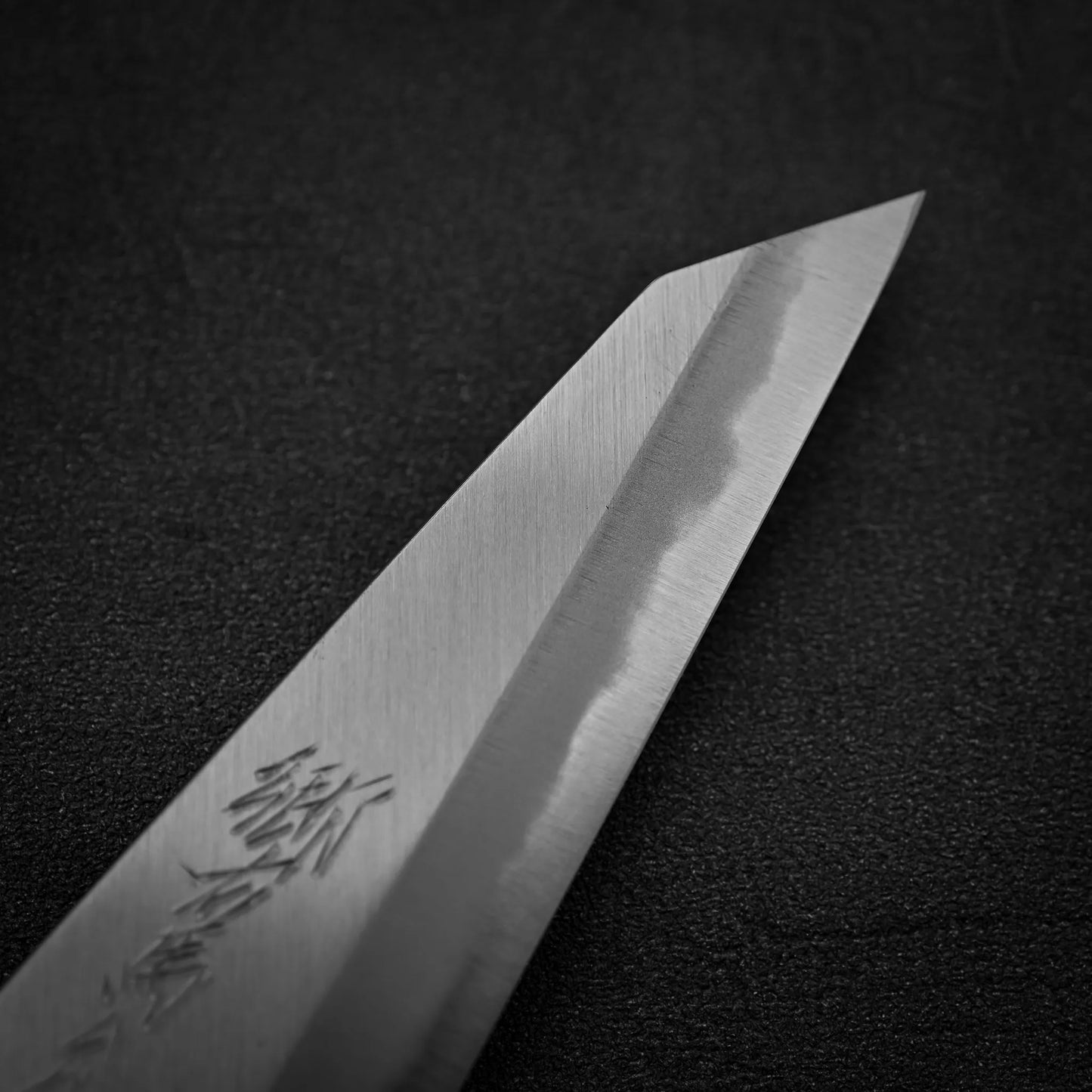 Close-up of the pointed tip of the Yoshikazu Tanaka shirogami#1 double bevel honesuki 170x50, showing detailed engraving and a unique shirogami#1 steel pattern on the blade, set against a dark textured background.