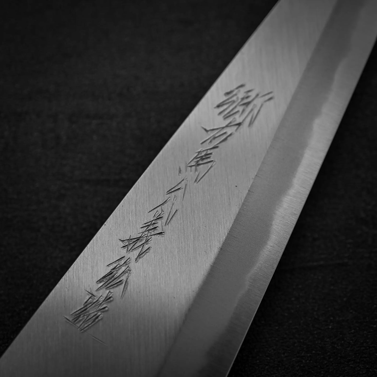A close-up of the Yoshikazu Tanaka shirogami#1 double bevel honesuki 170x50 shows its fine engravings on the polished blade, crafted by Yoshikazu Tanaka, with a razor-sharp edge set against a dark background.