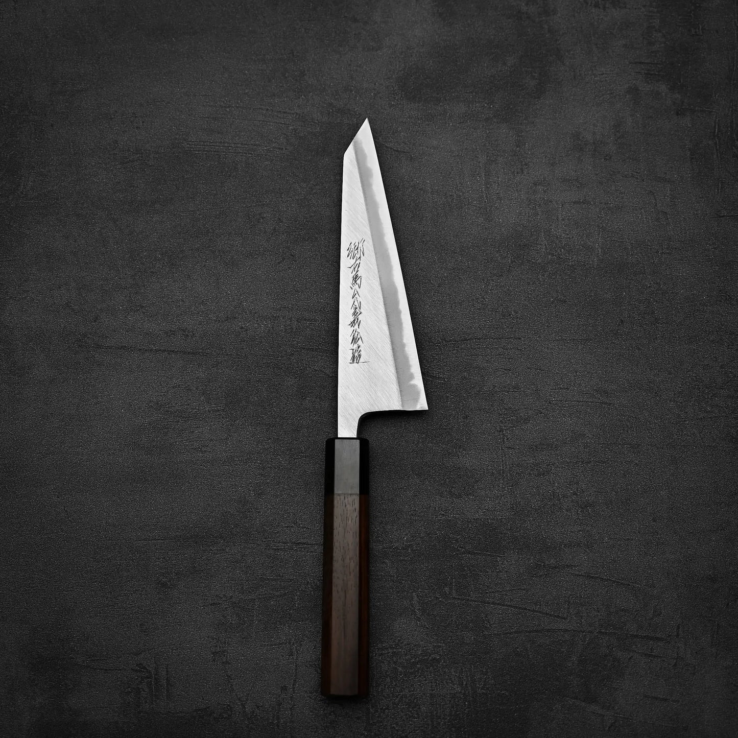 A Yoshikazu Tanaka shirogami#1 double bevel honesuki 170x50 with a sharp, angular blade and dark wooden handle rests on a textured surface; engraved Japanese characters decorate the blade.