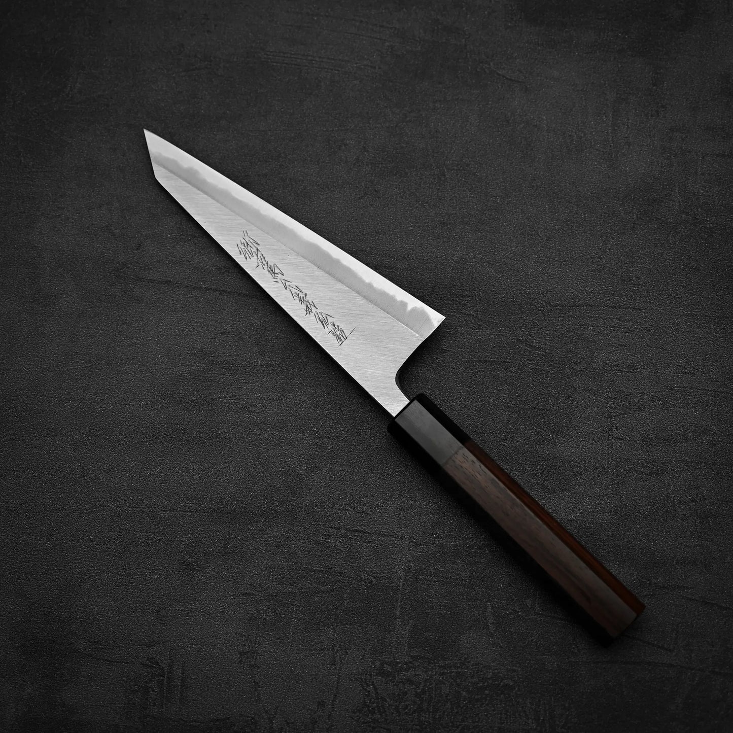 The Yoshikazu Tanaka shirogami#1 double bevel honesuki 170x50, crafted by Yoshikazu Tanaka with a pointed shirogami#1 steel blade and dark wooden handle engraved with Japanese characters, rests on a dark, textured surface.