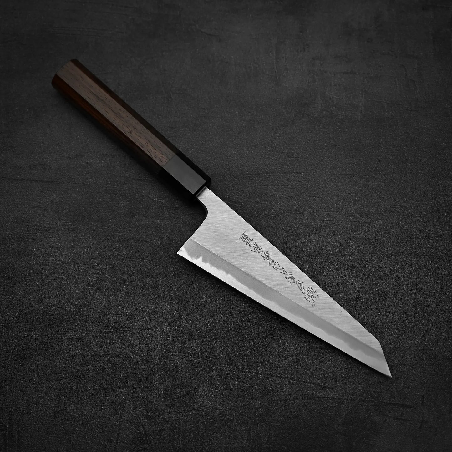 The Yoshikazu Tanaka shirogami#1 double bevel honesuki 170x50, featuring engraved Japanese characters and a wooden handle, rests on a dark textured surface.