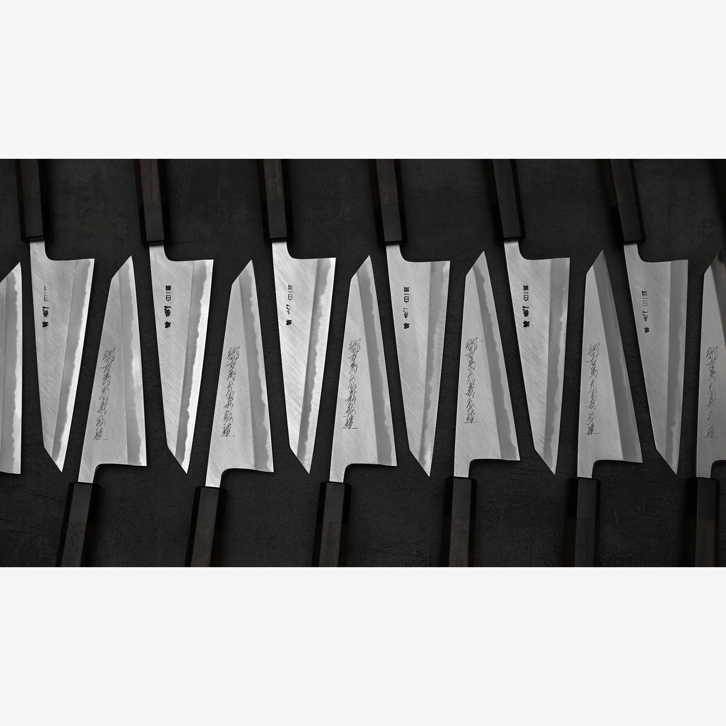 A Yoshikazu Tanaka shirogami#1 double bevel honesuki 170x50, along with other Japanese-style kitchen knives with wooden handles, is arranged in neat, angled rows on a dark surface. Each blade bears engraved characters.