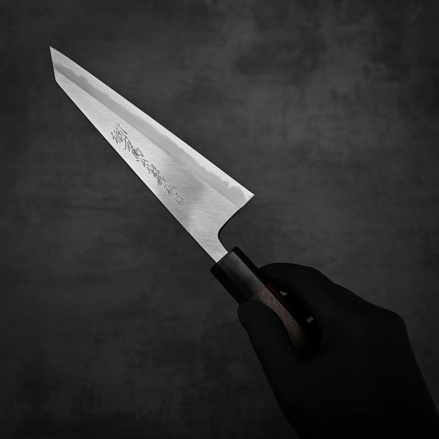 A hand holds the Yoshikazu Tanaka shirogami#1 double bevel honesuki 170x50, its pointed blade and wooden handle highlighted against a dark, textured background.