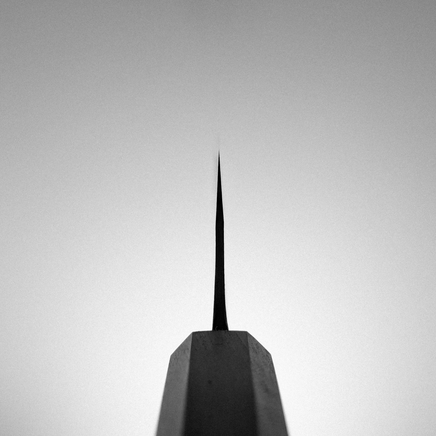A black and white photo shows the tip of the Kyohei Shindo kurouchi aogami#2 gyuto 210mm knife pointing upward, centered against a plain light background. The minimalist image is shot from a low angle.