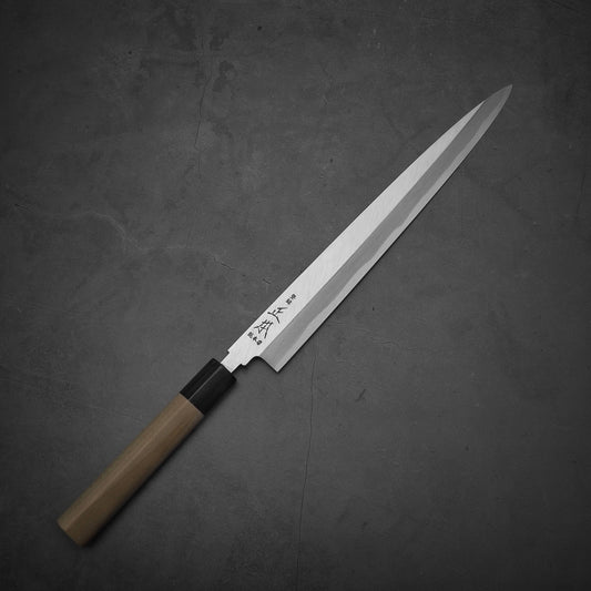 The Masamoto KS shirogami#2 yanagiba 270mm, a Japanese-style kitchen knife with a wooden handle and long, narrow shirogami#2 steel blade, features engraved Japanese characters near the handle and is displayed on a dark textured surface.