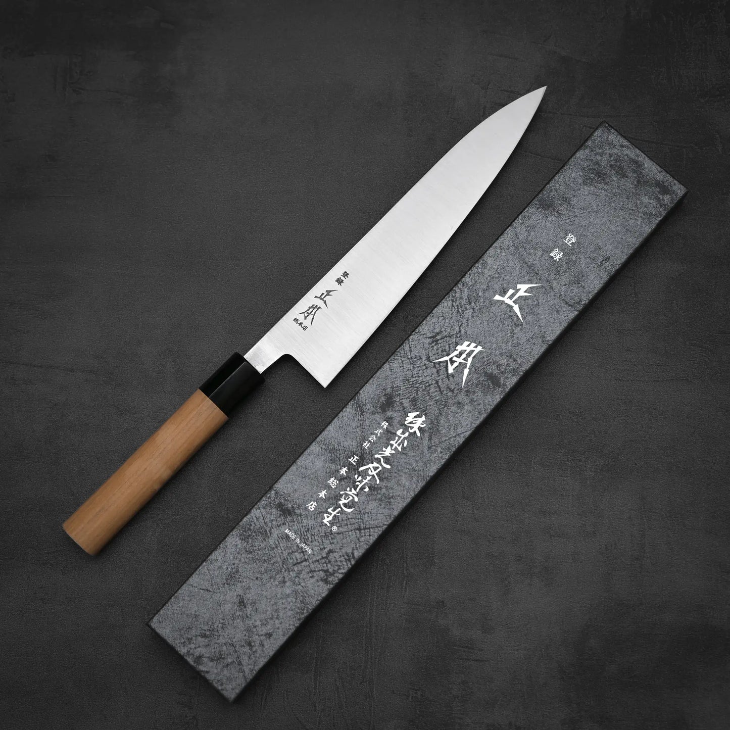 The Masamoto KS gyuto 210mm chef’s knife, featuring an ambidextrous wooden handle, lies diagonally beside its gray Masamoto box with Japanese lettering on the lid. Both rest on a dark textured surface.