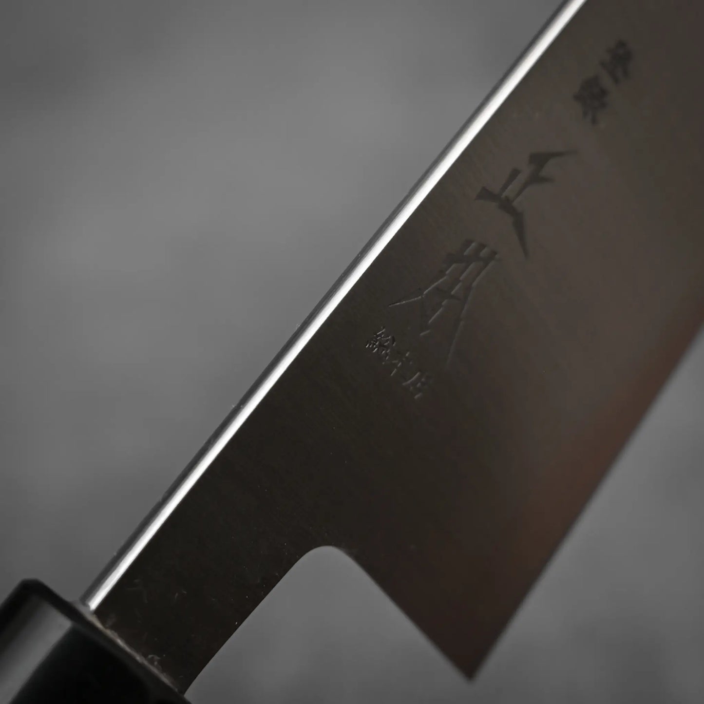 Close-up of the Masamoto KS gyuto 210mm blade made from Shirogami#2 steel, engraved with kanji, showcasing its sharp edge and part of the black handle by Masamoto against a blurred gray background.